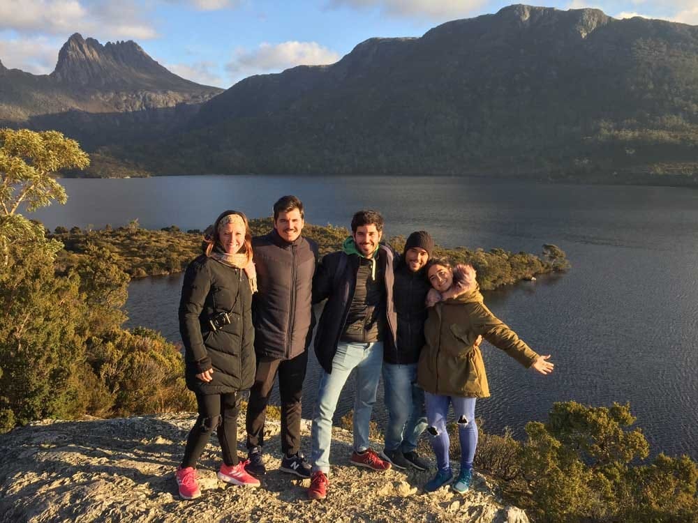 Vivir en Australia grupo de cuatro jóvenes sonriendo a cámara y detrás un lago y unas montañas